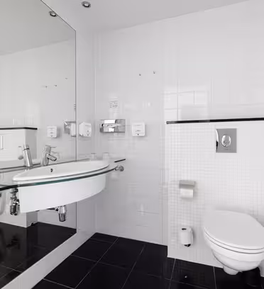 Et moderne badeværelse med hvide flisevægge og mørkt klinkegulv. Den har et vægmonteret toilet, en glashylde med en vask, et stort spejl og en bruser med glasdøre. Sæbedispensere monteres på væggen over vasken.