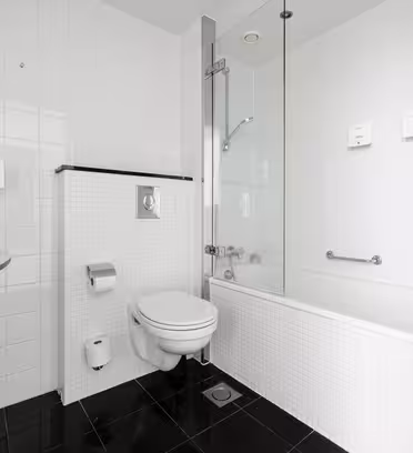 Moderne badeværelse med hvide flisevægge og sort klinkegulv. Funktionerne inkluderer en vægmonteret håndvask, toilet og et badekar med glasadskillelse og brusehoved. En toiletpapirholder støder op til toilettet.