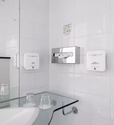 Et badeværelse med hvide fliser har en moderne glasvask med to plastikkopper. På væggen er sæbe- og lotiondispensere monteret sammen med en servietholder. Et glas brusekabine er synligt i baggrunden.