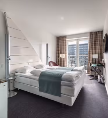 Et moderne hotelværelse med en stor seng med hvidt sengetøj og et blågrønt tæppe. Et skrivebord med en stol er tæt på vinduet, som har udsigt over et bylandskab. Et væghængt tv og en minibar med drinks og snacks er også synlige.