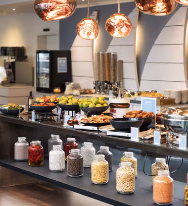 En moderne buffetopstilling med et udvalg af frisk frugt, korn og forskellige fødevarer udstillet i krukker og fade. Ovenpå hænger pendullamper af kobber, og rammerne har et slankt, moderne design med kaffemaskine og køleskab i baggrunden.