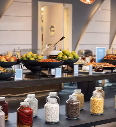Et buffetbord i moderne rammer, med skåle med æbler, pærer, kager og diverse snacks. Nedenfor vises krukker med ingredienser som pasta og korn. Baggrunden viser en lækker indretning med blød belysning.