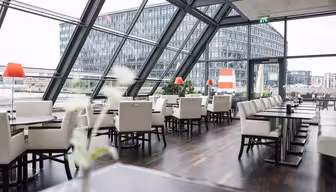 Moderne restaurantinteriør med vinklede glasvægge, der afslører en udsigt over byen. Rummet har hvide polstrede stole, træborde og røde lampeaccenter. Store vinduer giver udsigt til en kontorbygning i baggrunden på en overskyet dag.