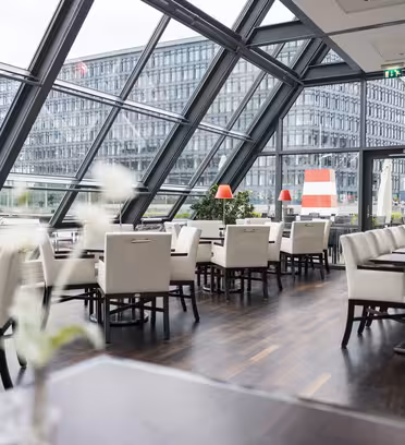 Moderne restaurantinteriør med vinklede glasvægge, der afslører en udsigt over byen. Rummet har hvide polstrede stole, træborde og røde lampeaccenter. Store vinduer giver udsigt til en kontorbygning i baggrunden på en overskyet dag.