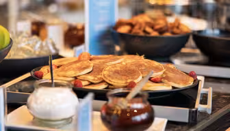 Et bord med en buffet med et stort fad pandekager pyntet med jordbær. I nærheden er skåle med flødeskum og sirup, med andre morgenmadsprodukter sløret i baggrunden.