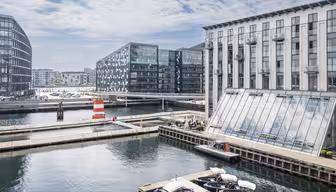 Moderne havnescene med slanke kontorbygninger ved vandet, inklusive en karakteristisk trekantet a-rammestruktur. Bådene ligger til kaj i forgrunden. Et lille rød-hvidstribet fyrtårn står på en mole under en delvist overskyet himmel.