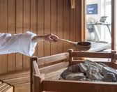 En person i en hvid morgenkåbe hælder vand fra en slev på varme saunasten inde i et saunarum af træ. Fitnessudstyr er synligt i baggrunden.