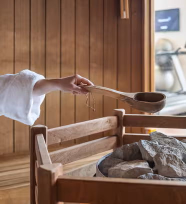 En person i en hvid morgenkåbe hælder vand fra en slev på varme saunasten inde i et saunarum af træ. Fitnessudstyr er synligt i baggrunden.