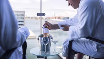 To personer i hvide badekåber sidder på en balkon med udsigt over vandet. En person holder en flaske rosévin og hælder den i et glas på et lille rundt bord med et andet glas og en isspand. Udsigten omfatter moderne bygninger i det fjerne.