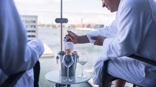 To personer i hvide badekåber sidder på en balkon med udsigt over vandet. En person holder en flaske rosévin og hælder den i et glas på et lille rundt bord med et andet glas og en isspand. Udsigten omfatter moderne bygninger i det fjerne.