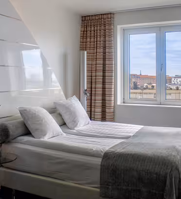 Et moderne hotelværelse med en dobbeltseng, hvidt sengetøj, fire puder, en rød stol og et stort vindue med stribede gardiner med udsigt til et bybillede under en blå himmel.