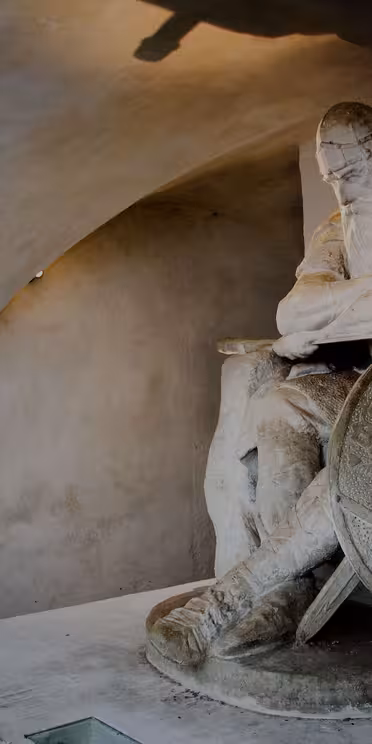 En stenstatue af en siddende kriger med et langt skæg, hjelm og en kappe, hvilende en arm på et stort skjold, der er indviklet dekoreret med cirkulære mønstre. Statuen er placeret i en svagt oplyst buet alkove.