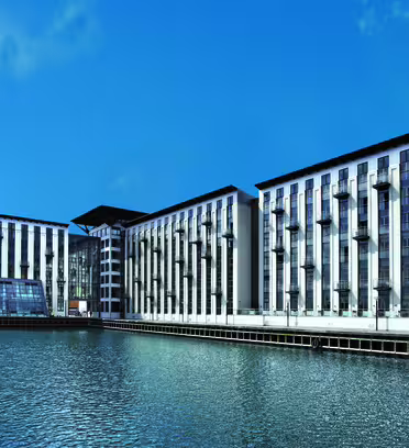 Moderne bygning ved vandet med høje, lodrette vinduer og altaner ud mod et vandområde. Strukturen har et symmetrisk design og er sat mod en klar blå himmel.