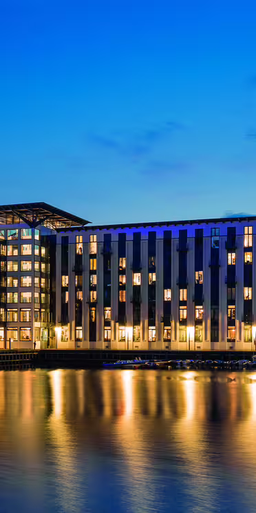 Moderne bygning med store vinduer, oplyst om natten, reflekterer på roligt vand. Strukturen har en blanding af lodrette og vandrette linjer i sin arkitektur. Himlen er dybblå, hvilket indikerer tusmørke.