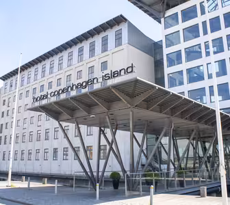 Moderne bygning af Hotel Copenhagen Island med minimalistisk design. Funktionerne omfatter en glasindgang understøttet af metalbjælker og store vinduer. Hotellets navn er tydeligt vist nær indgangen.