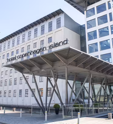 Moderne bygning af Hotel Copenhagen Island med minimalistisk design. Funktionerne omfatter en glasindgang understøttet af metalbjælker og store vinduer. Hotellets navn er tydeligt vist nær indgangen.