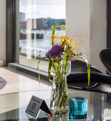 En glasvase med farverige blomster står på et rundt glasbord ved siden af en blå lysestage. I baggrunden har moderne sorte stole og store vinduer udsigt over et bybillede og vandmasse.