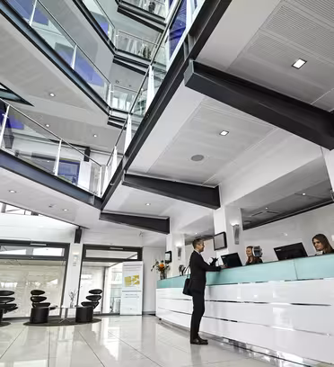 En moderne kontorlobby med en elegant reception, hvor en person taler med to receptionister. Rummet har store vinduer, flere etager og moderne indretning med hvide og sorte accenter.