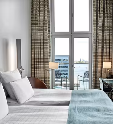 Moderne hotelværelse med dobbeltseng prydet med hvidt og aqua-sengetøj, ud mod et stort vindue med stribede gardiner. Vinduet giver udsigt til en havnefront. Et vægmonteret tv og et glasbord er på siden.