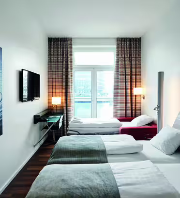 Moderne hotelværelse med dobbeltseng og enkeltseng. Værelset har et stort vindue med udsigt over byen, et vægmonteret tv og et moderne kunstværk med vandtema. Indretningen omfatter hvide vægge og mørkt trægulv.