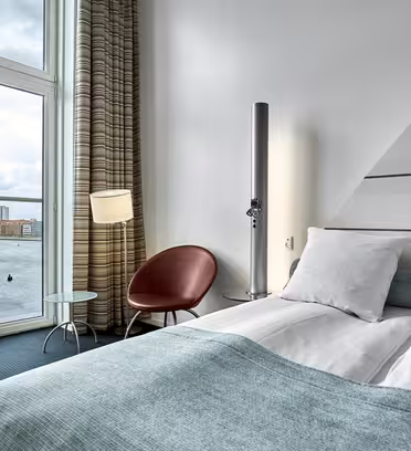Moderne hotelværelse med stort vindue med udsigt over vandet. Værelset har en pæn opredt seng med et lyseblåt og hvidt farveskema, en lækker stålampe, en rund stol og et lille rundt glasbord. Neutraltonede gardiner hænger ved vinduet.