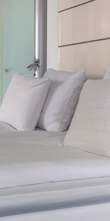 Moderne hotelværelse med en pænt redt seng med hvidt sengetøj og flere puder. En træhylde ovenover rummer forskellige ting, og en glasdør står lidt på klem i baggrunden, hvilket tilføjer det slanke, minimalistiske design.