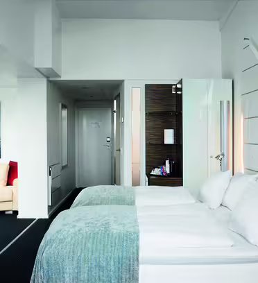 Moderne hotelværelse med to pænt opredte enkeltsenge, hvidt sengetøj og lyseblå sengetøj. Et fladskærms-tv er monteret på venstre væg. Der er et lille loungeområde med en sofa og farverige puder. Værelset har minimal indretning og et slankt, rent design.