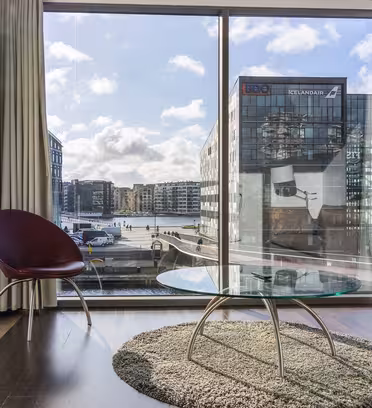 En moderne stue med et stort glasvindue med udsigt over bybygninger og en vandvej. Indeni er der et rundt sofabord i glas på et tæppe, en rødbrun stol og en beige sofa. Sollys strømmer ind og skaber en lys og luftig atmosfære.