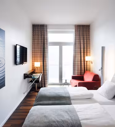 Et moderne hotelværelse med to senge, hver med hvidt sengetøj og grønne sengetøj. En rød lænestol sidder ved et vindue med gennemsigtige, ternede gardiner. Der er et tv monteret på væggen og et stort indrammet billede, der viser vandbølger.