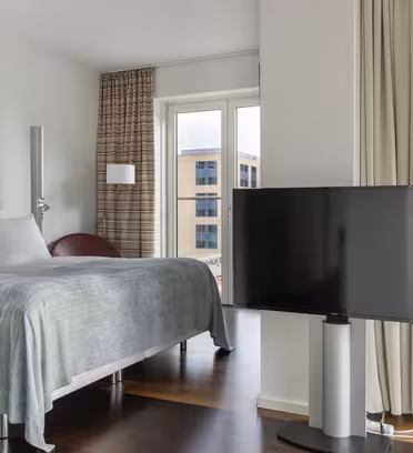 Moderne hotelværelse med en pænt redt seng, accentstol og fladskærms-tv. Store vinduespartier giver udsigt til bybilledet og vandveje med lysgardiner delvist fortrukket. Rummet har et rent, minimalistisk design med neutrale toner.