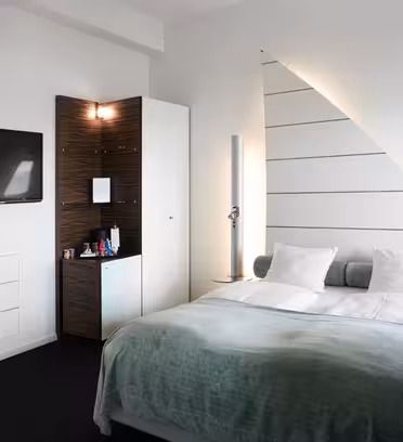 Moderne hotelværelse med dobbeltseng dækket af et lysegrønt tæppe og hvide puder. Der er et vægmonteret fladskærms-tv, en trekantet hjørneenhed med bekvemmeligheder og stilfulde høje cylindriske højttalere ved siden af sengen. Væggene er hvide og slanke.
