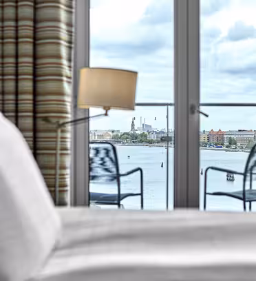 Et hyggeligt værelse med en seng i forgrunden og udsigt til et bybillede gennem en glasdør. Balkonen udenfor har to stole og har udsigt over et vandområde. En lampe er placeret nær døren, med en overskyet himmel synlig bagved.