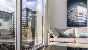 En hyggelig, moderne stue med en beige sofa, blå hynder og et cirkulært maleri med krusningseffekt. Store vinduer viser et bybillede med høje bygninger, herunder en mærket "HANDELSBANKEN." Et glas sofabord fuldender rammerne.