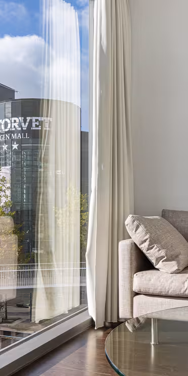 En hyggelig, moderne stue med en beige sofa, blå hynder og et cirkulært maleri med krusningseffekt. Store vinduer viser et bybillede med høje bygninger, herunder en mærket "HANDELSBANKEN." Et glas sofabord fuldender rammerne.