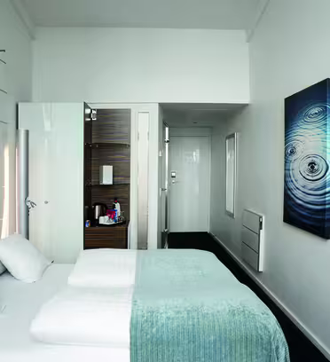Et moderne hotelværelse med en pænt redt seng med hvidt sengetøj og et blågrønt tæppe. En dør fører til et lille badeværelse. Væggene er hvide, prydet med et abstrakt kunstværk, og der er monteret et fladskærms-tv på den ene væg.