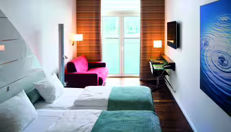 Moderne hotelværelse med to senge med blågrønne tæpper og hvide puder. En rød lænestol sidder ved et vindue med stribede gardiner. Et væghængt tv og skrivebord med en stol er til højre, og et stort kunstværk med vand ripple design er på væggen.
