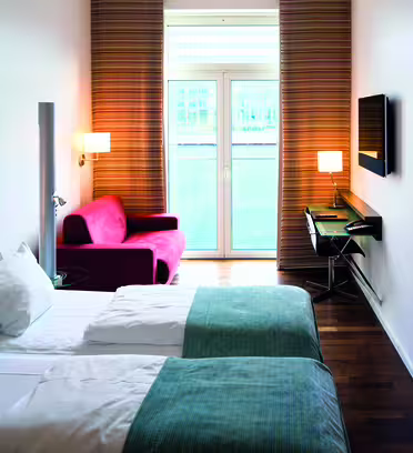Moderne hotelværelse med to senge med blågrønne tæpper og hvide puder. En rød lænestol sidder ved et vindue med stribede gardiner. Et væghængt tv og skrivebord med en stol er til højre, og et stort kunstværk med vand ripple design er på væggen.
