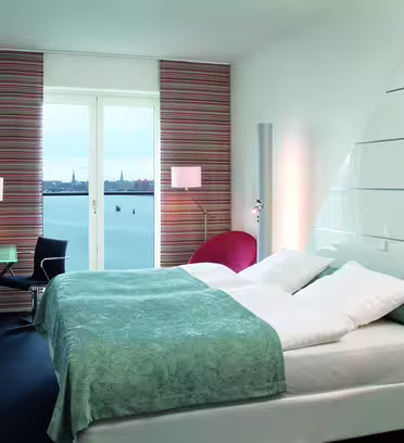 Moderne hotelværelse med stor seng klædt i hvidt og lyseblåt sengetøj. Der er et vinduesudsigt over et roligt vandområde og et fjernt bybillede. Værelset har et elegant skrivebord, stol og en rød lænestol med minimalistisk indretning.