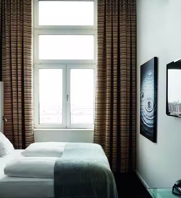 Et moderne hotelværelse har en pænt redt seng med hvidt og gråt sengetøj, et stort vindue med mønstrede gardiner, et vægmonteret fladskærms-tv og abstrakt kunst. En telefon hviler på et bord med glasplade. Naturligt lys fylder rummet.