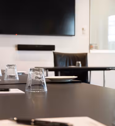 Et konferencelokale har et stort tv på væggen, et langbord med briller, kuglepenne og papirer på hovedet. En stol er synlig for bordet, med en tallerken og et glas på hovedet foran.