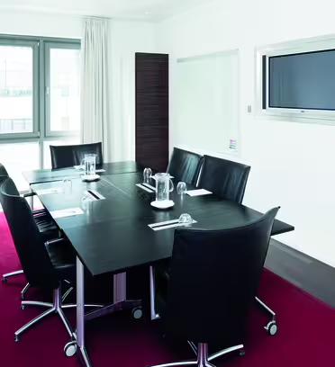 Et moderne mødelokale med et sort bord og seks sorte læderstole på en rød løber. Rummet har et stort vindue, en vægmonteret skærm og whiteboards på hver side. Glas og vandkander stilles på bordet.