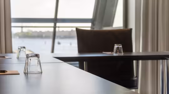 Der vises et moderne mødelokale med borde og vendte glas. Store vinduer giver udsigt til en vandmasse og fjerne bygninger. Lysgardiner indrammer scenen, og et blødt omgivende lys fylder rummet.