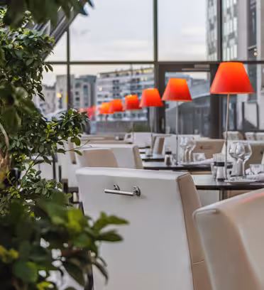 Moderne restaurant med hvide stole og borde til spisning. Røde lamper står langs bordene og tilføjer en varm glød. Store vinduer giver udsigt til byens bygninger. Indendørs planter delvist synlige i forgrunden.