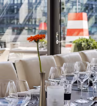 Elegant spisestue med hvide borddækninger og glasvarer på et langt, mørkt bord. To orange gerbera-margueritter i vaser tilføjer et glimt af farve. Store vinduer afslører en urban baggrund med en rød og hvid stribet struktur synlig udenfor.