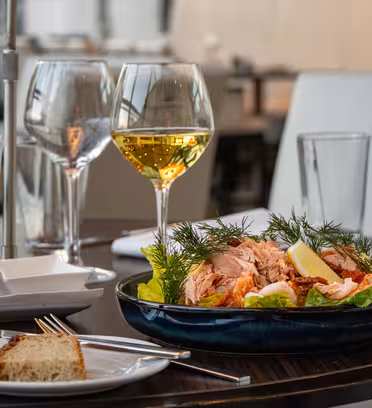 Et bord med en tallerken salat toppet med laks, citronskiver og dild, to glas hvidvin og en lille tallerken brød. Baggrunden har en moderne spisestue med blød belysning.
