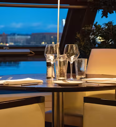 Et svagt oplyst restaurantbord til fire med hvide servietter, sølvbestik og tomme vinglas. Baggrunden viser et stort vindue med en sløret udsigt over byens skyline i skumringen. To polstrede stole er synlige.