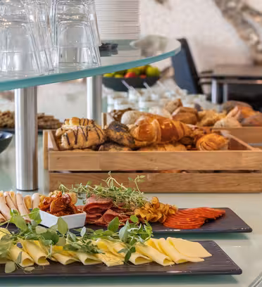 Et buffetbord med et fad med diverse oste, salami og krydderurter i forgrunden. Bagved er der kurve med diverse brød og kager. Briller er stablet til venstre, og et siddeområde er synligt i baggrunden.