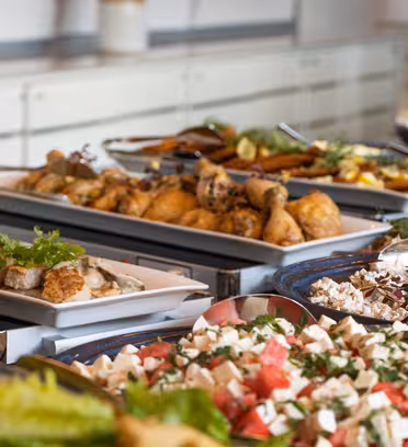 Et buffetbord fyldt med forskellige retter, herunder salater, ristede kyllingeunderlår, pasta og pyntet kød. Frisk grønt og krydderurter bruges som pynt. Tallerkener og redskaber er synlige i baggrunden.