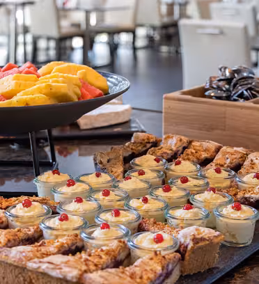 Et buffetbord med et udvalg af desserter, herunder små glaskopper med cremede desserter toppet med røde bær, diverse skiver smuldrede kager og en skål med blandede frugtskiver som vandmelon og ananas, anbragt i en restaurant.