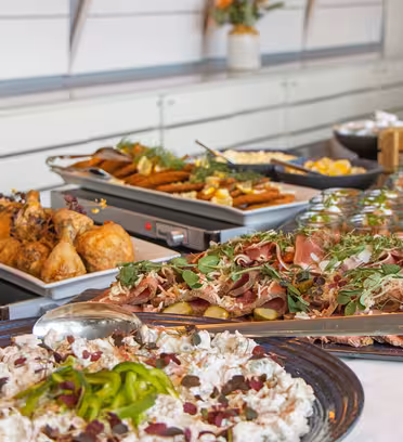 Et buffetbord med forskellige retter, herunder stegt kylling, kød i skiver med grønt, grillede grøntsager og salater. Tallerkener med mad er smukt arrangeret, klar til gæster. I baggrunden fremhæver dekorative blomster stemningen.